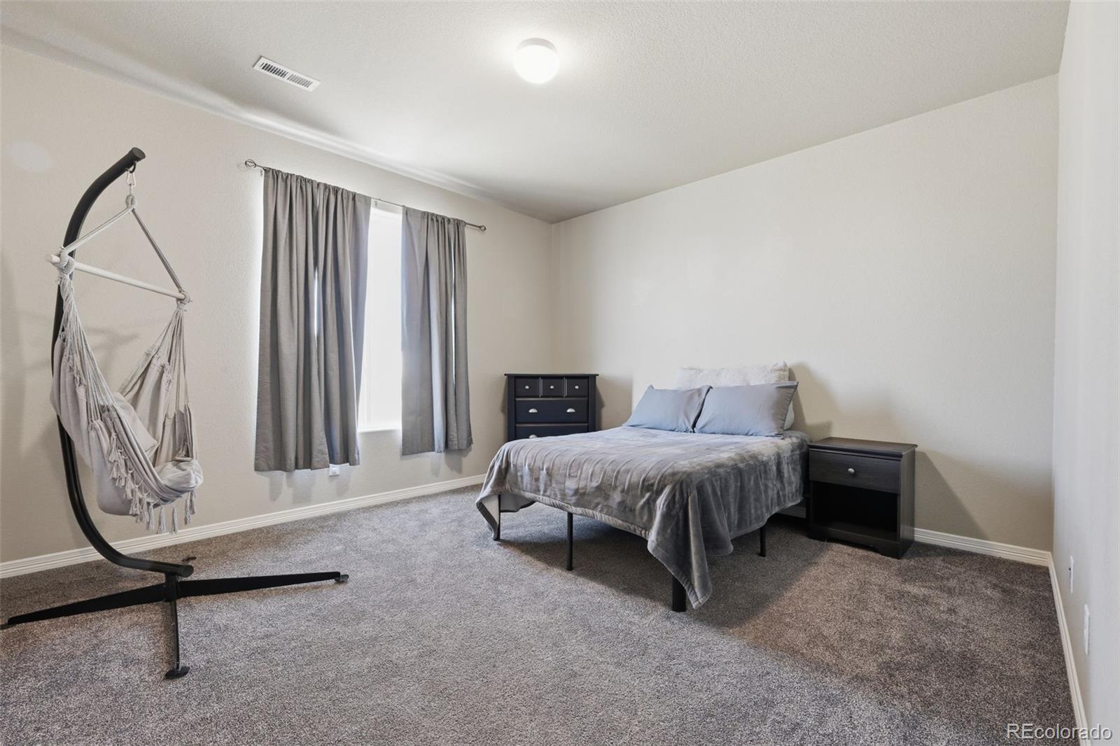Furnished bedroom with a gray-covered bed, dark nightstands, and a hanging chair beside the window.