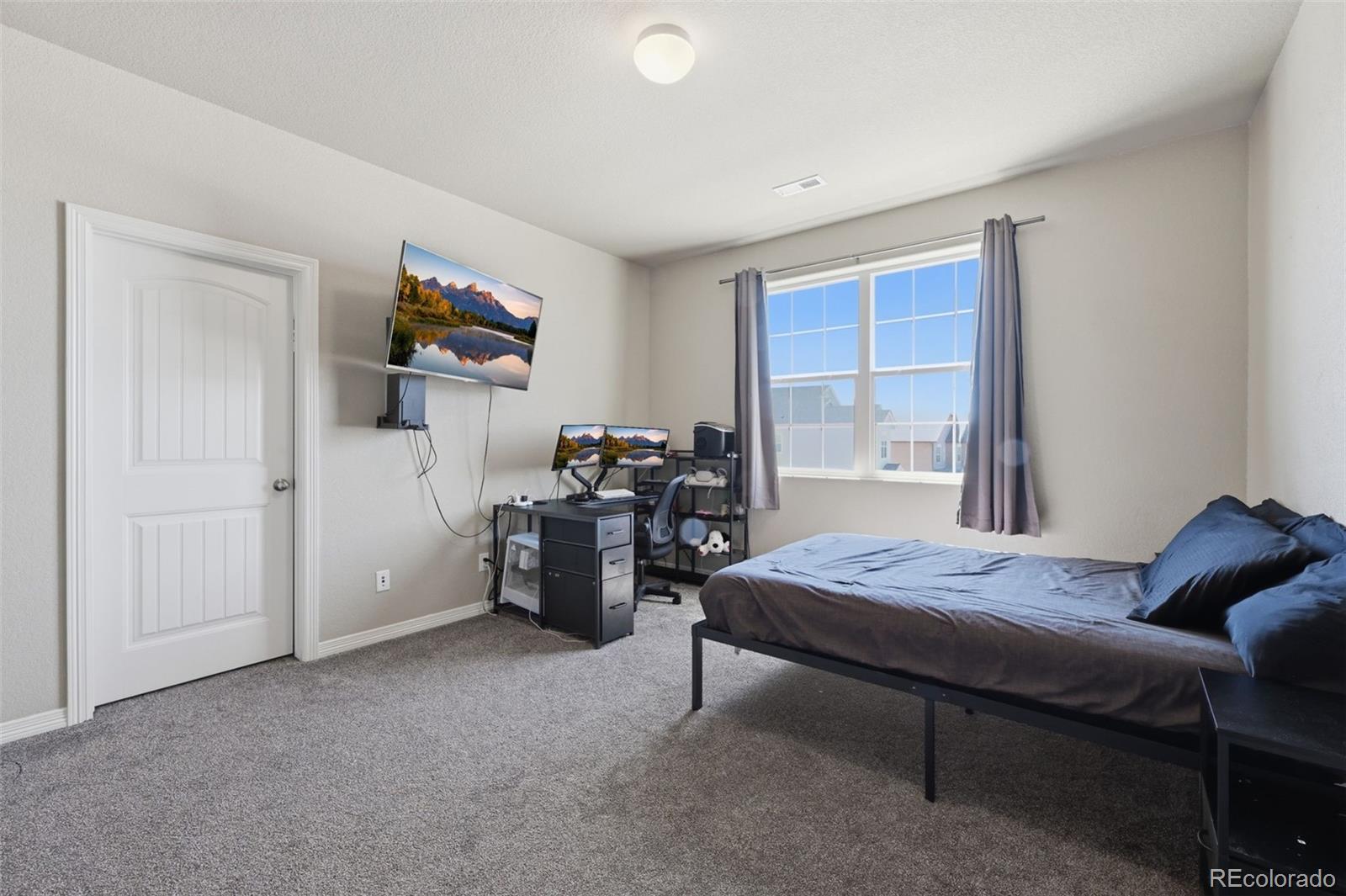 Bedroom with a computer desk and monitors beside a bed with blue pillows.