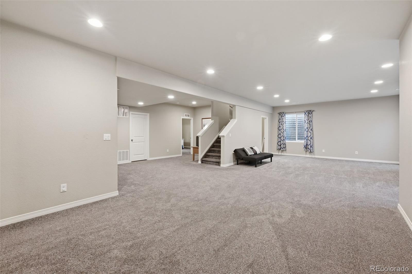 Finished basement living area with broad gray carpet, recessed lights, and a staircase.