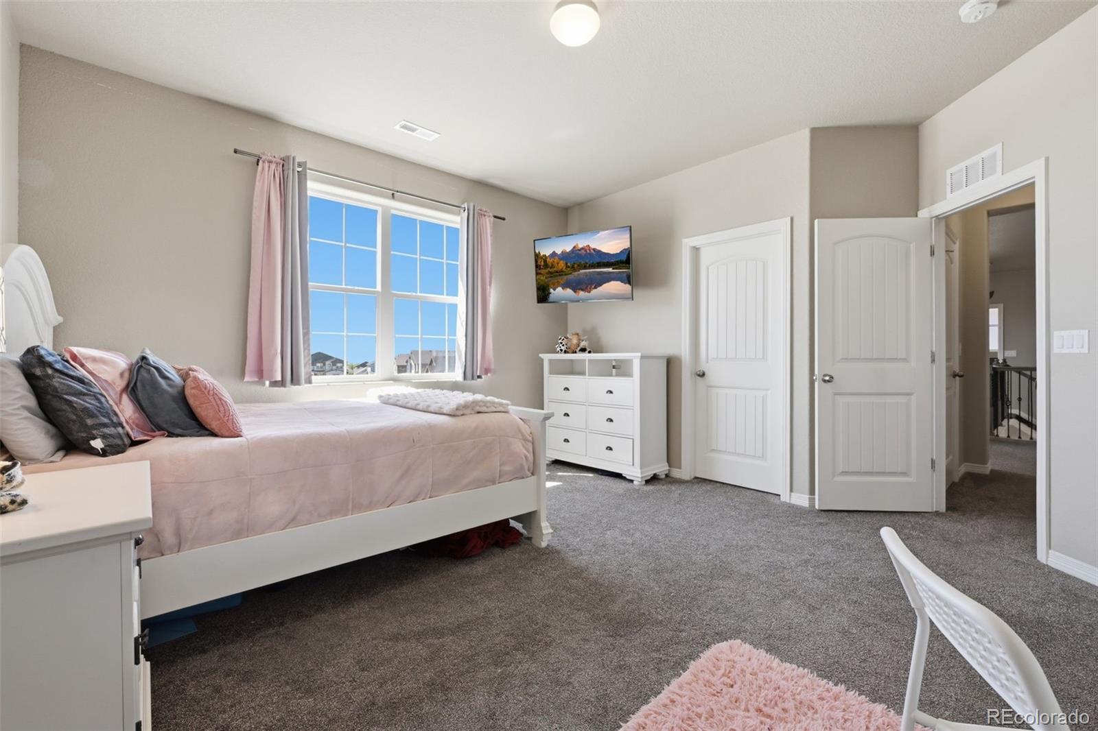 Furnished bedroom with a pink bedspread, gray carpet, and a large window with pink curtains.