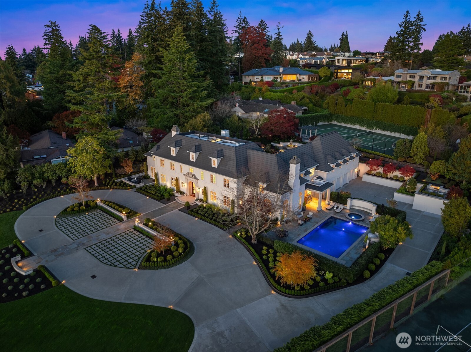 Aerial view of a large white estate with a circular driveway, swimming pool, and tennis court at dusk.