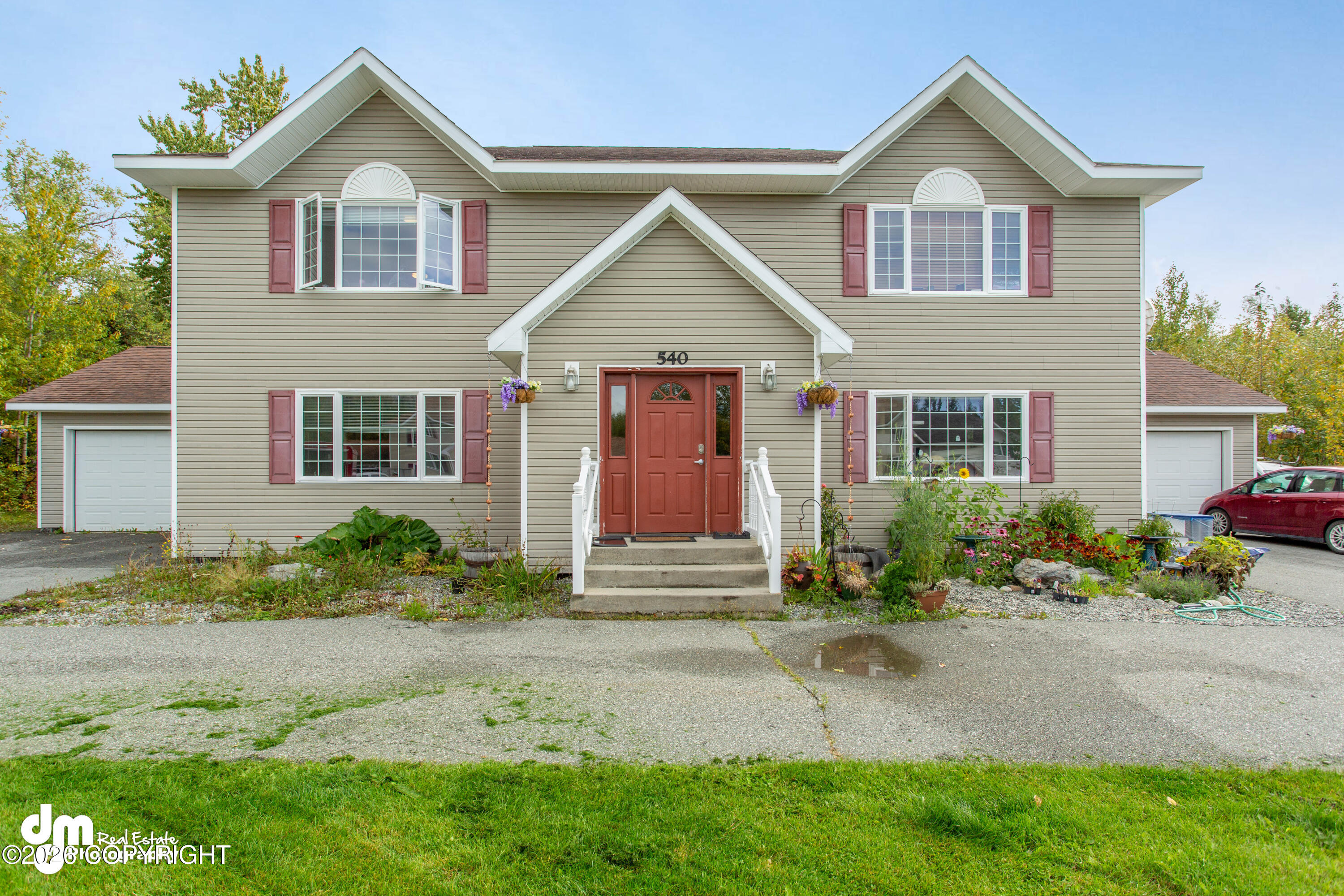 Two-story beige house with red shutters, a red front door, attached garages, and colorful garden beds.