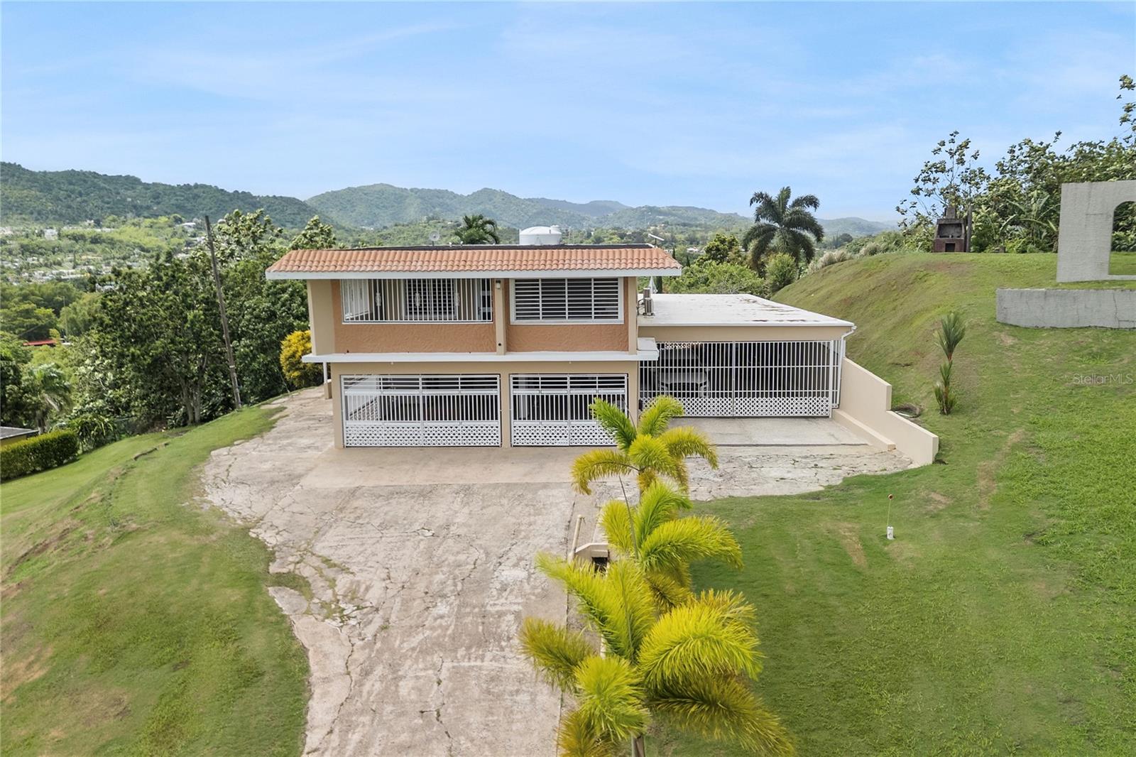 Two-story home with a tile roof and covered carport, set on a sloping palm-lined lot with mountain views.