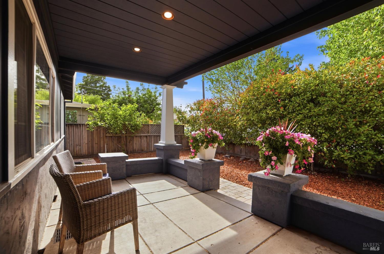 Covered backyard patio with wicker chairs, flowering planters, and a wooden privacy fence.