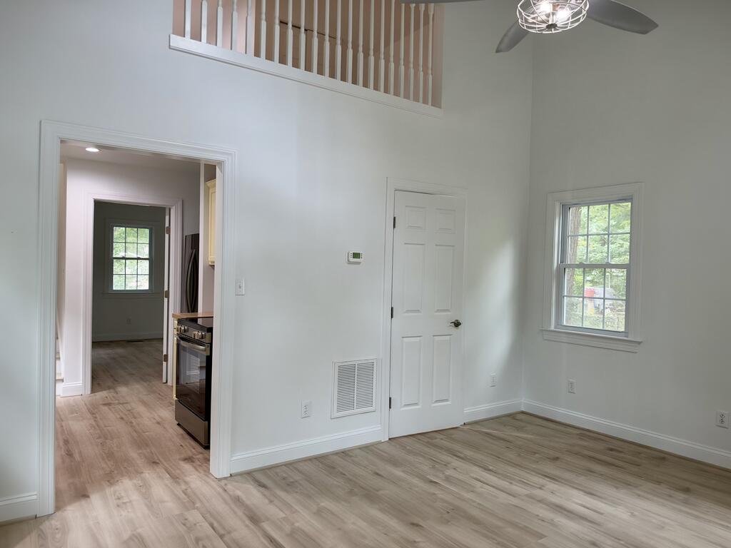 Vacant room with light wood floors, a ceiling fan, loft railing, and kitchen visible through the doorway.