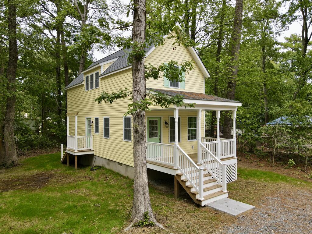 Pale yellow two-story house with a covered white-railed porch among mature trees.