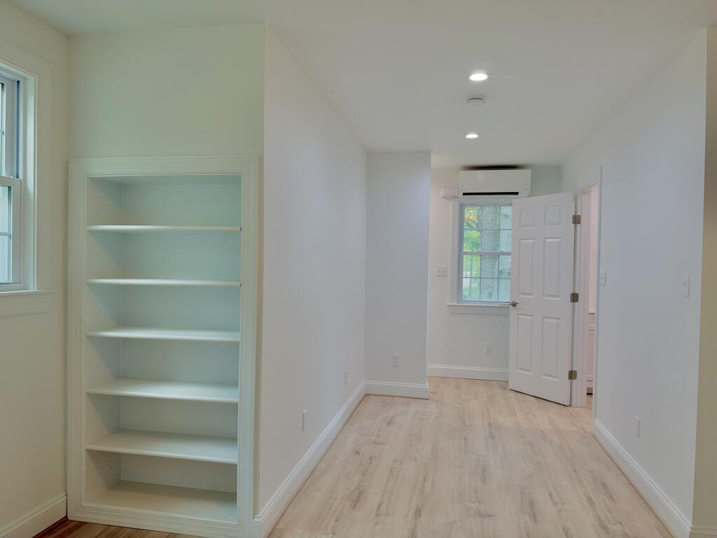 Empty hallway with built-in shelves, pale wood flooring, and a window beside an open door.