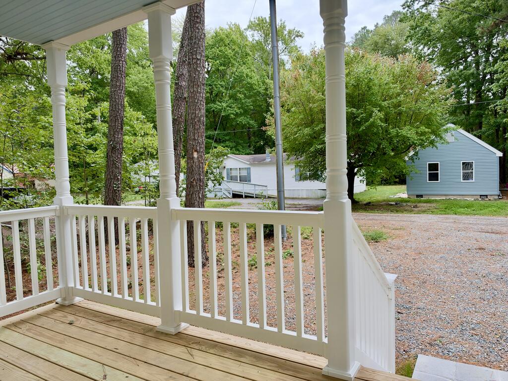 Covered porch with white railings overlooks a gravel driveway, neighboring houses, and mature trees.