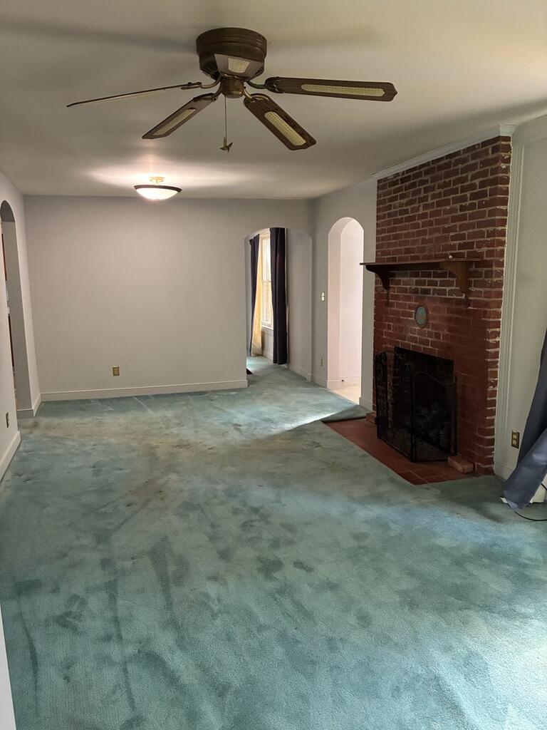Unfurnished living room with teal carpet, a brick fireplace, and a ceiling fan.
