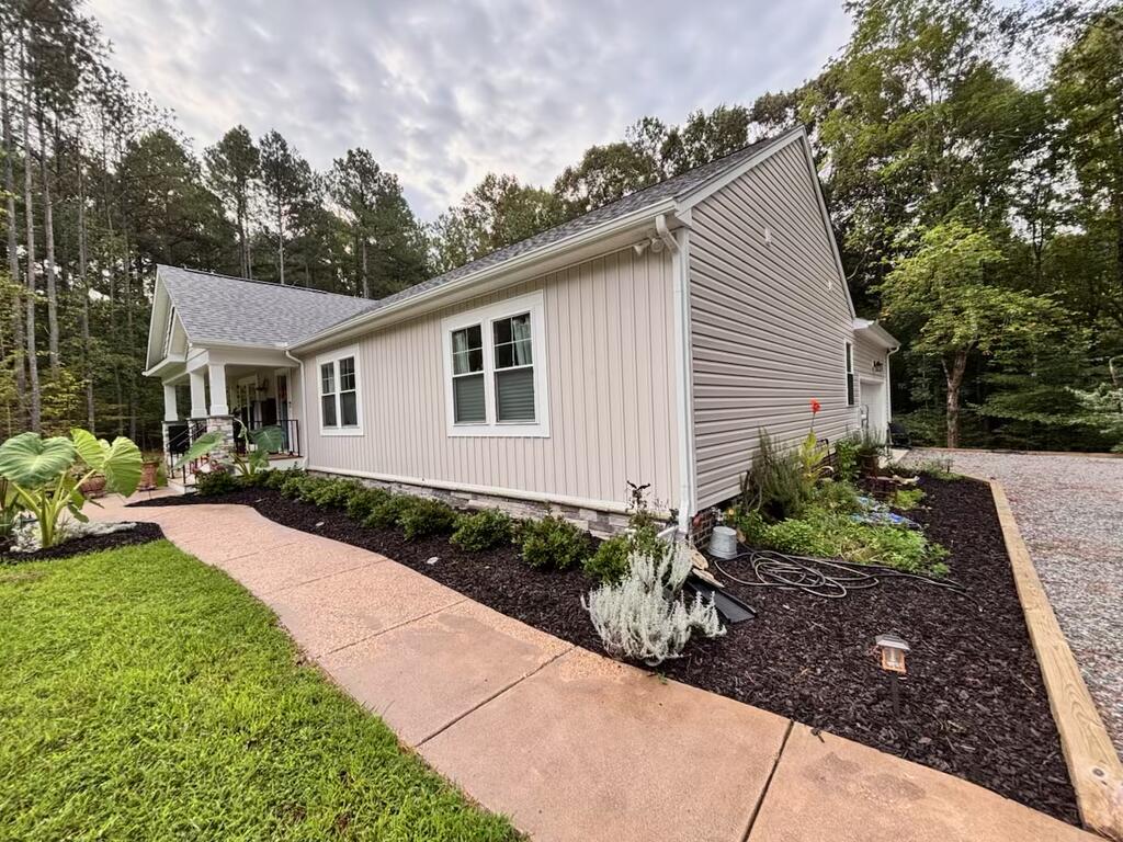 Light-colored home with a covered porch, landscaped beds, and a curved walkway, surrounded by mature trees.