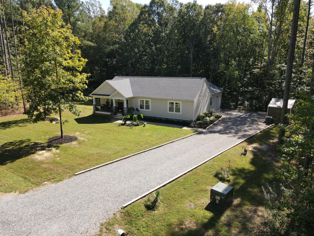Single-story home with a long gravel driveway, lawn, and mature trees surrounding the property.