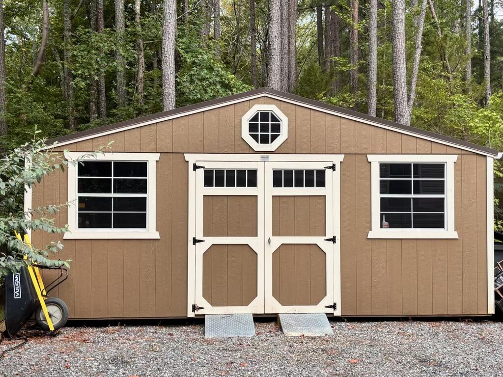 Tan storage shed with white trim, double doors, and an octagonal window, surrounded by trees.