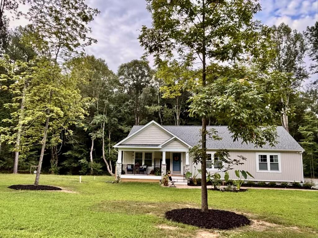 White single-story home with a covered porch, broad lawn, and wooded backdrop.