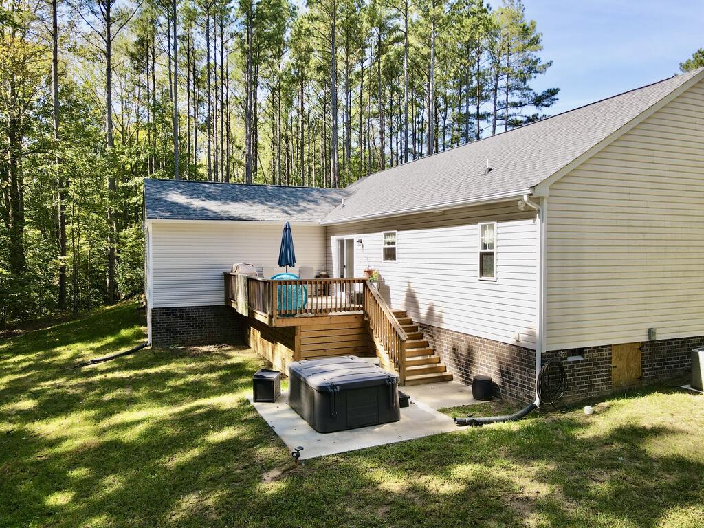 Rear of a light-sided home with a raised deck, stairs, and hot tub in a wooded backyard.