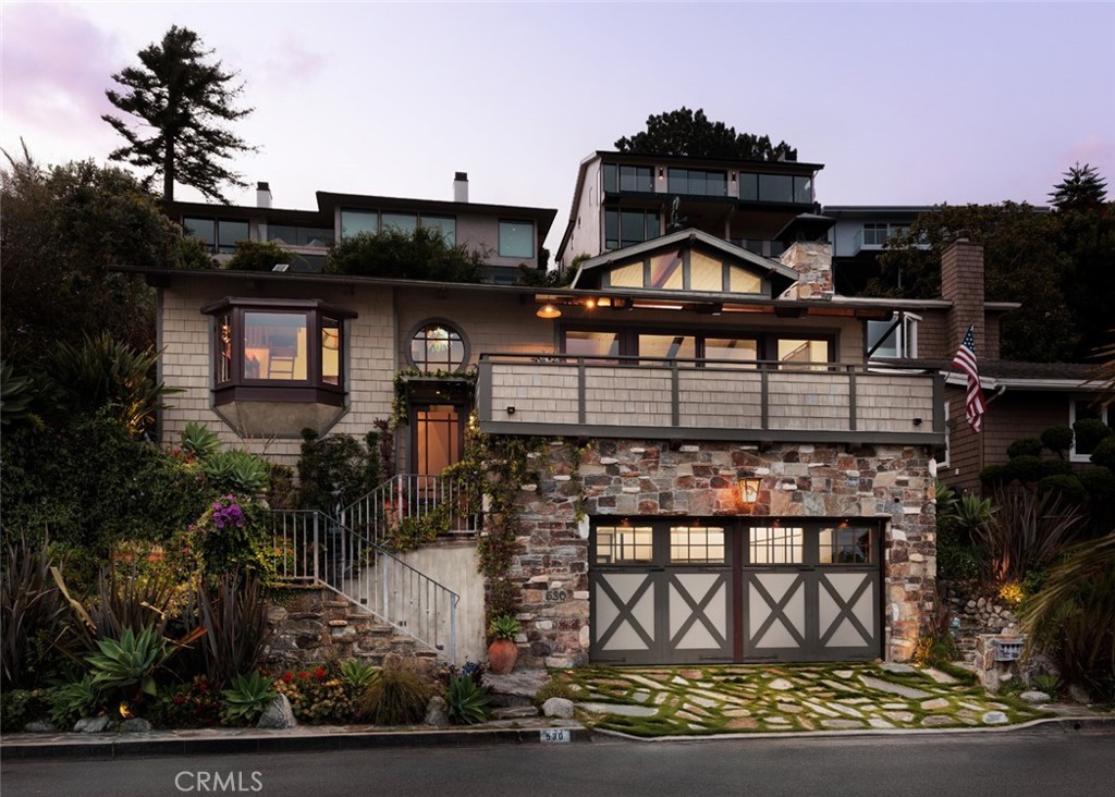 Multi-level home with stone facade, broad balcony, cross-braced garage doors, and landscaped approach at dusk.