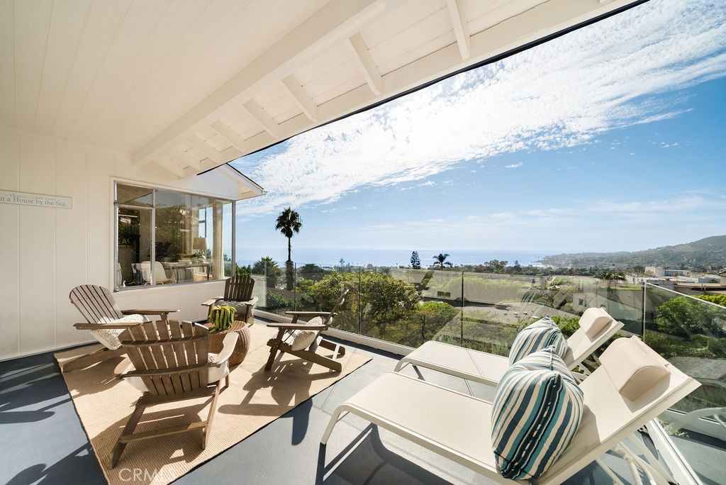 Covered terrace with lounge chairs and a glass railing overlooking the ocean and coastal hills.
