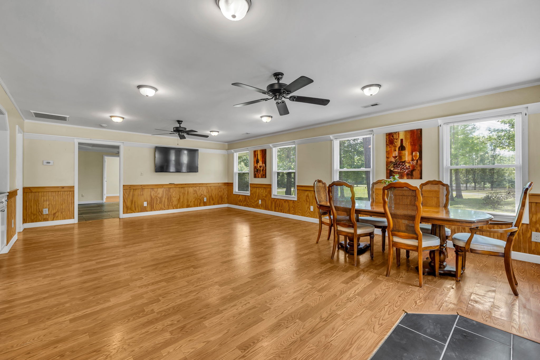 Open dining and living area with hardwood floors, wood paneling, large windows, and two ceiling fans.