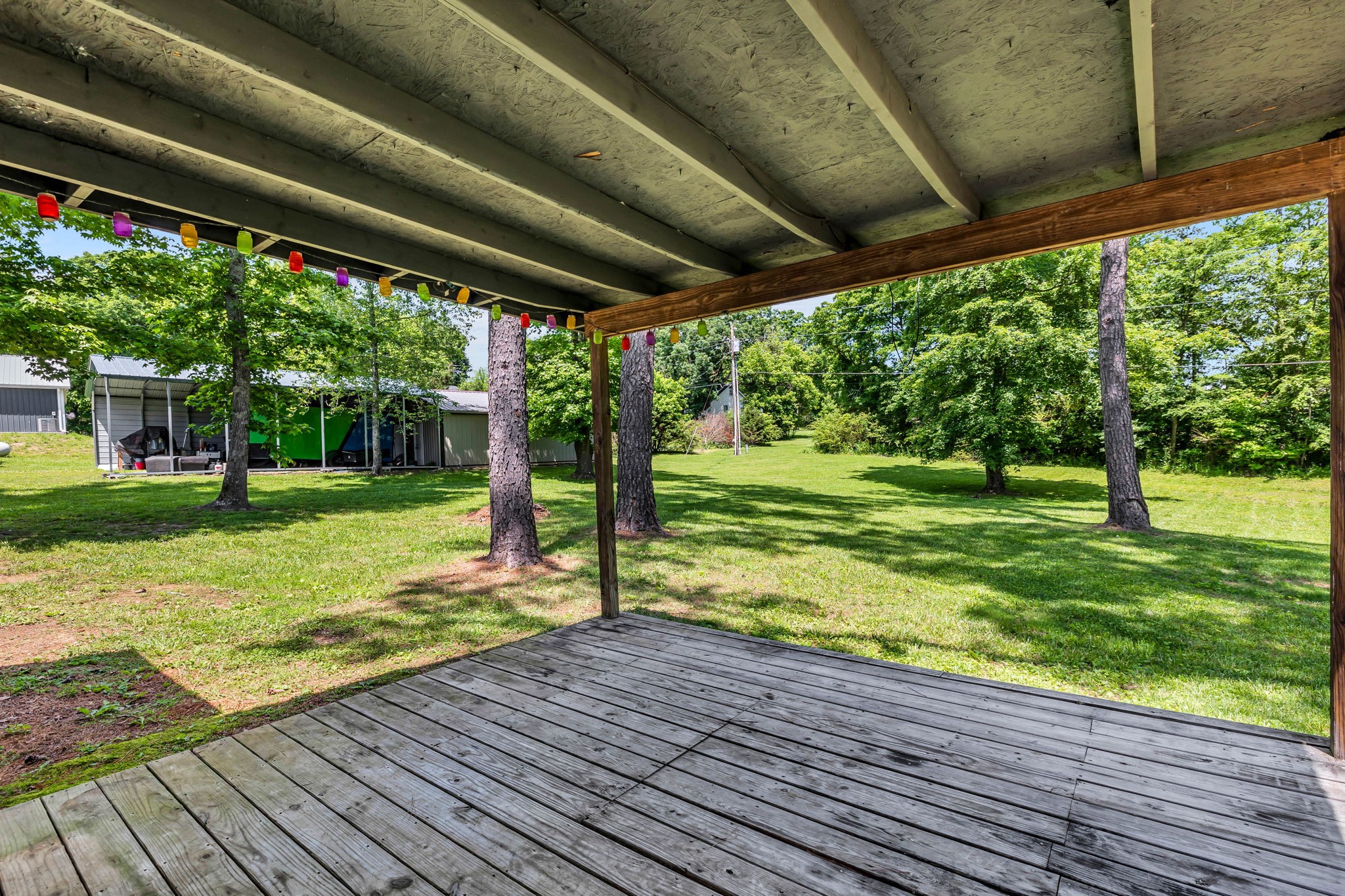 Covered wooden deck with colorful string lights overlooking a tree-shaded grassy yard.