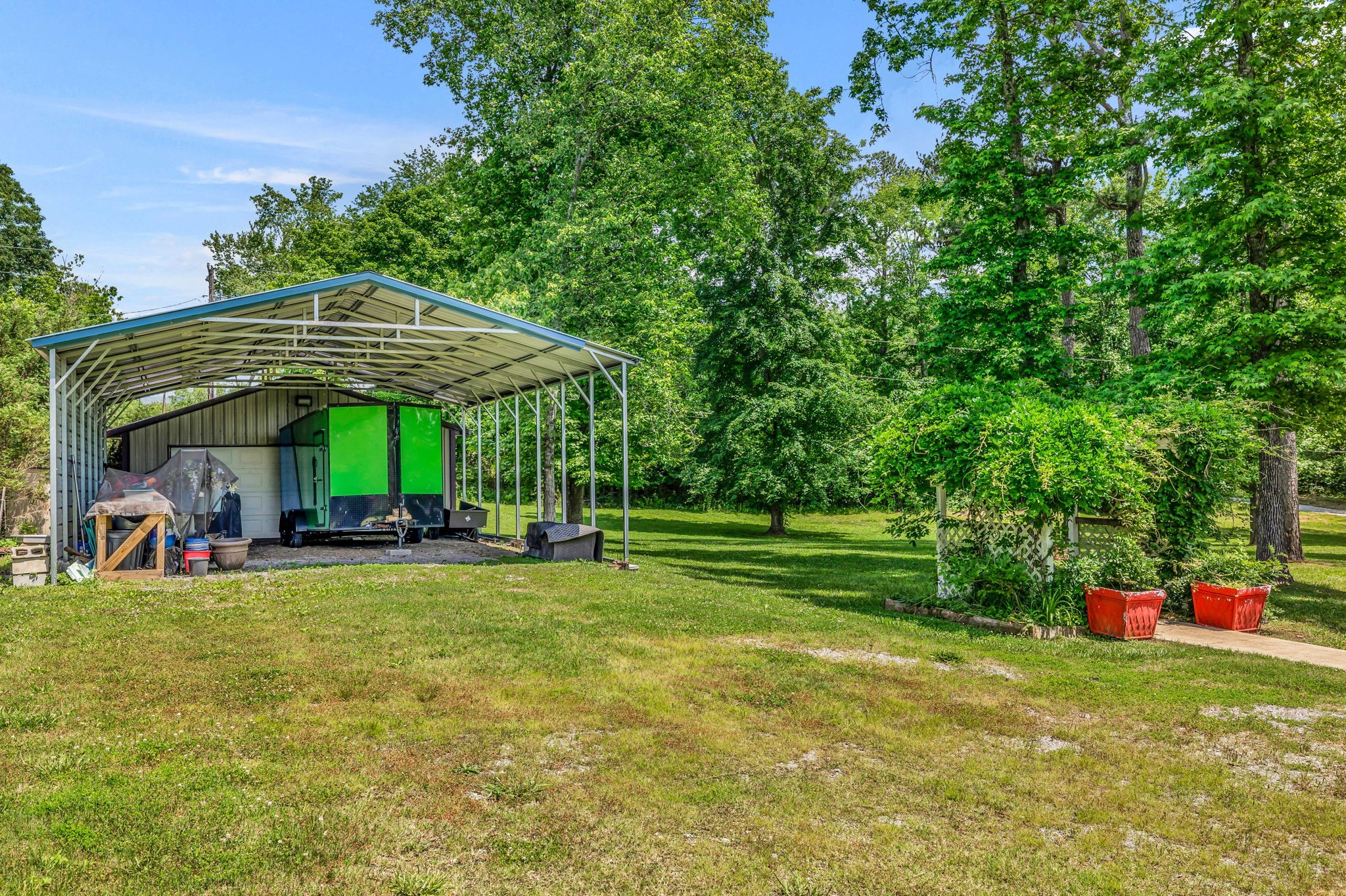 Tree-lined yard with a metal carport sheltering an outbuilding and green trailer.