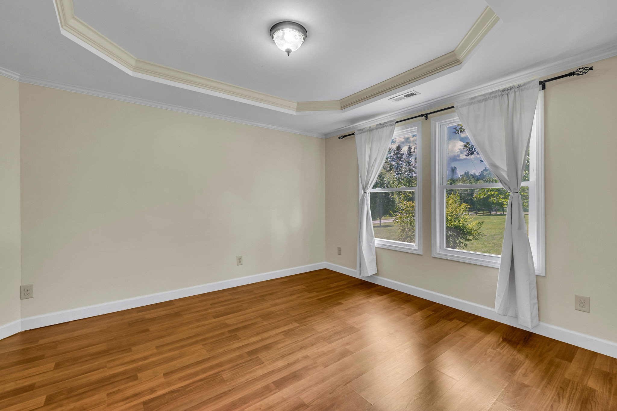 Empty room with wood floors, a tray ceiling, and curtained windows overlooking a green lawn.