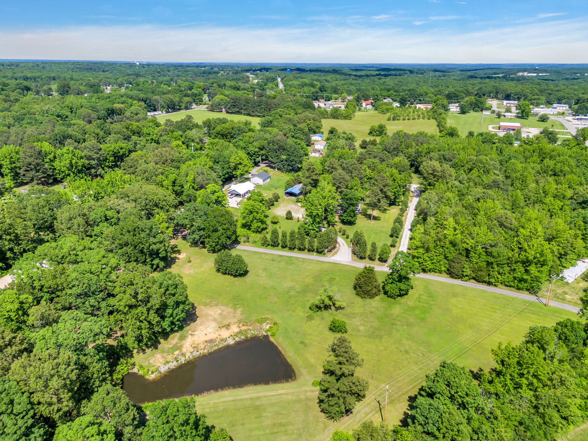 Aerial view of a wooded property with a pond, open lawn, and several buildings.