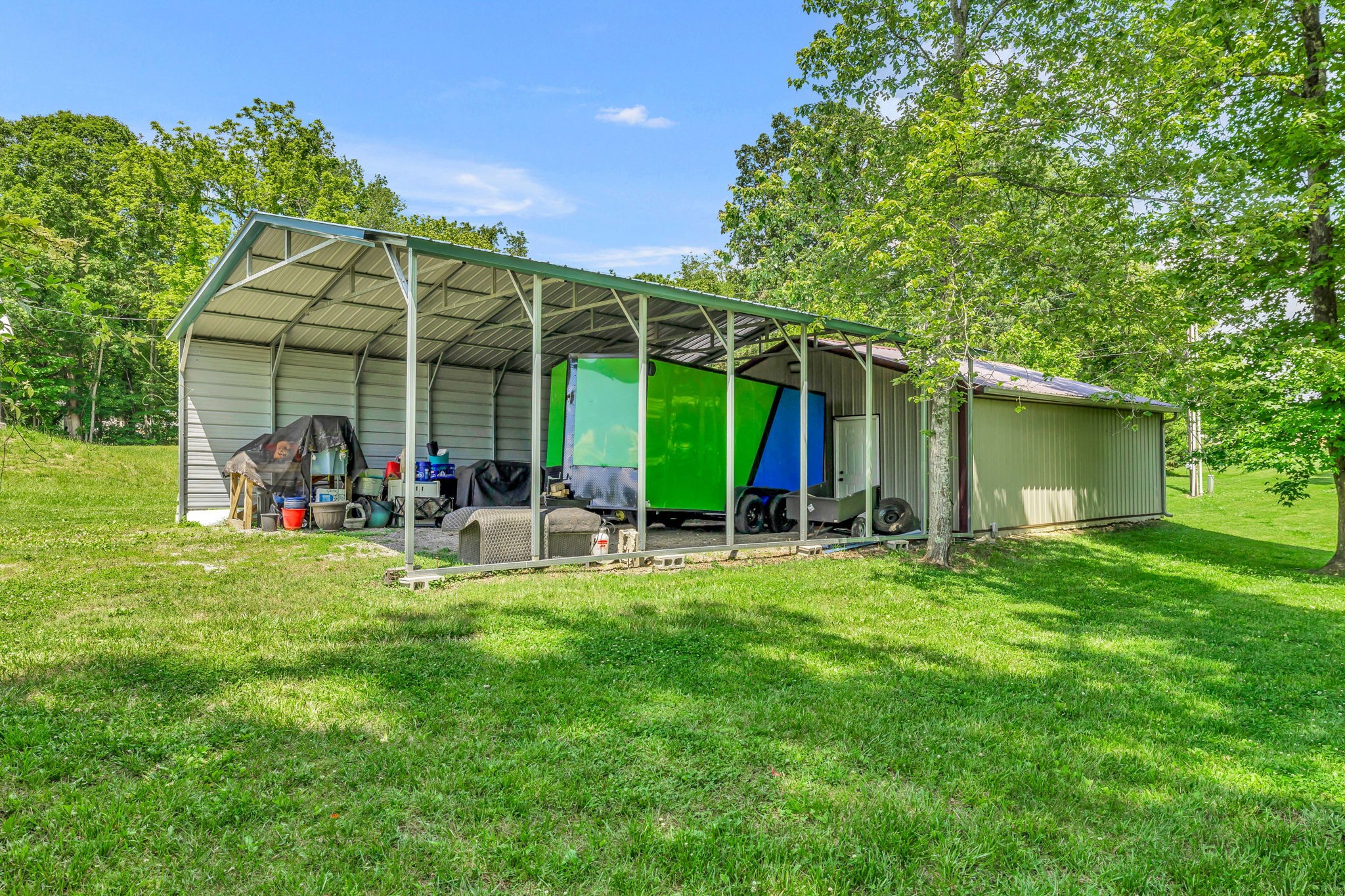 Open-sided metal carport shelters colorful trailers beside an outbuilding in a grassy, wooded yard.