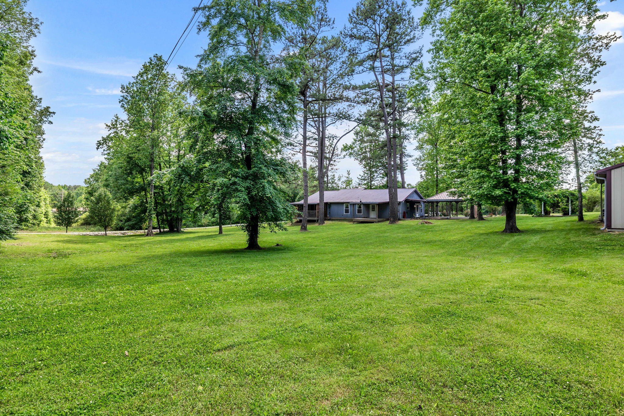 Single-story home beyond a broad lawn, surrounded by mature trees, with a metal roof and covered carport.