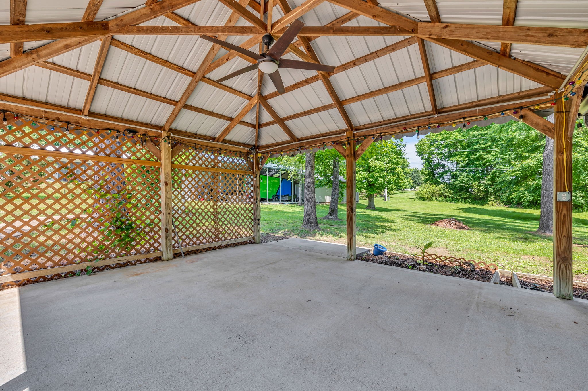 Covered concrete patio with exposed wooden trusses, a ceiling fan, and lattice overlooking a leafy yard.