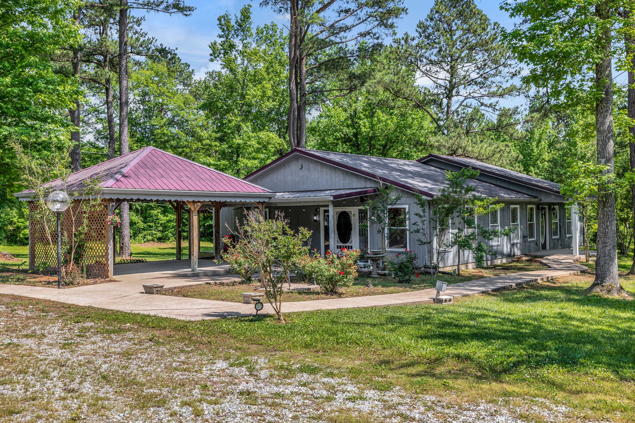 Gray single-story home among trees, with a burgundy-roofed covered carport and curved walkway.
