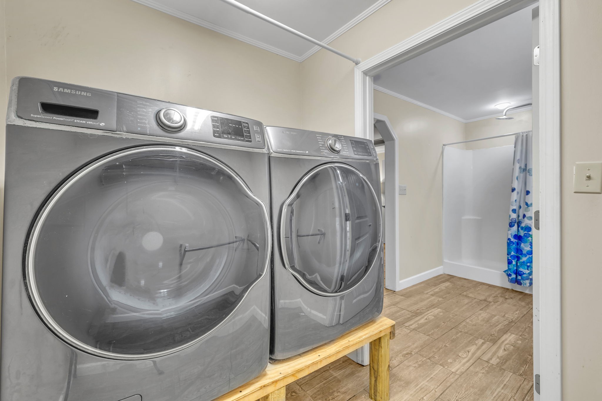 Laundry room with gray front-loading washer and dryer beside a bathroom with a blue-patterned shower curtain.