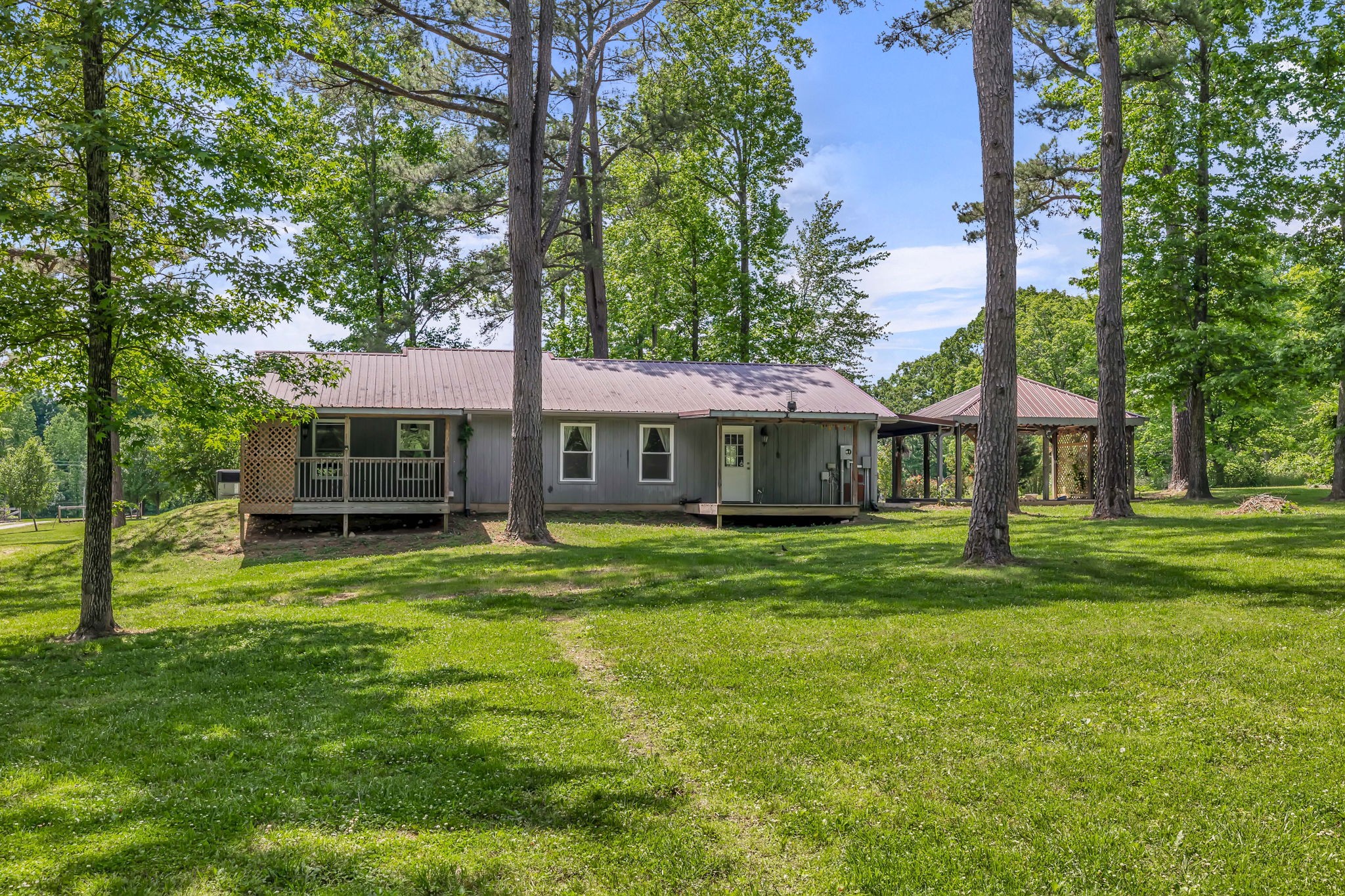 Single-story gray home with a metal roof, lattice porch, and open-sided pavilion among mature trees.