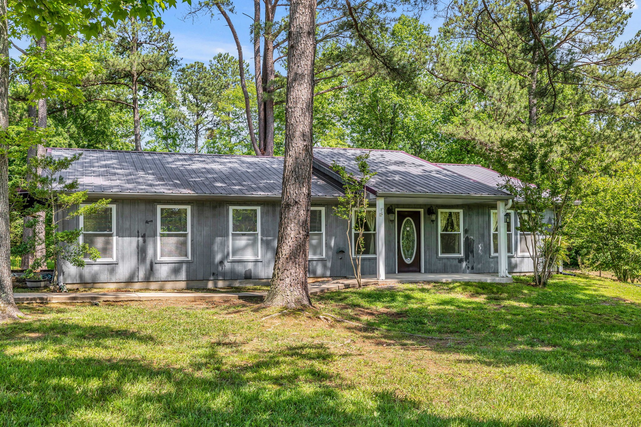 Gray single-story home with a metal roof and covered entry among mature trees.