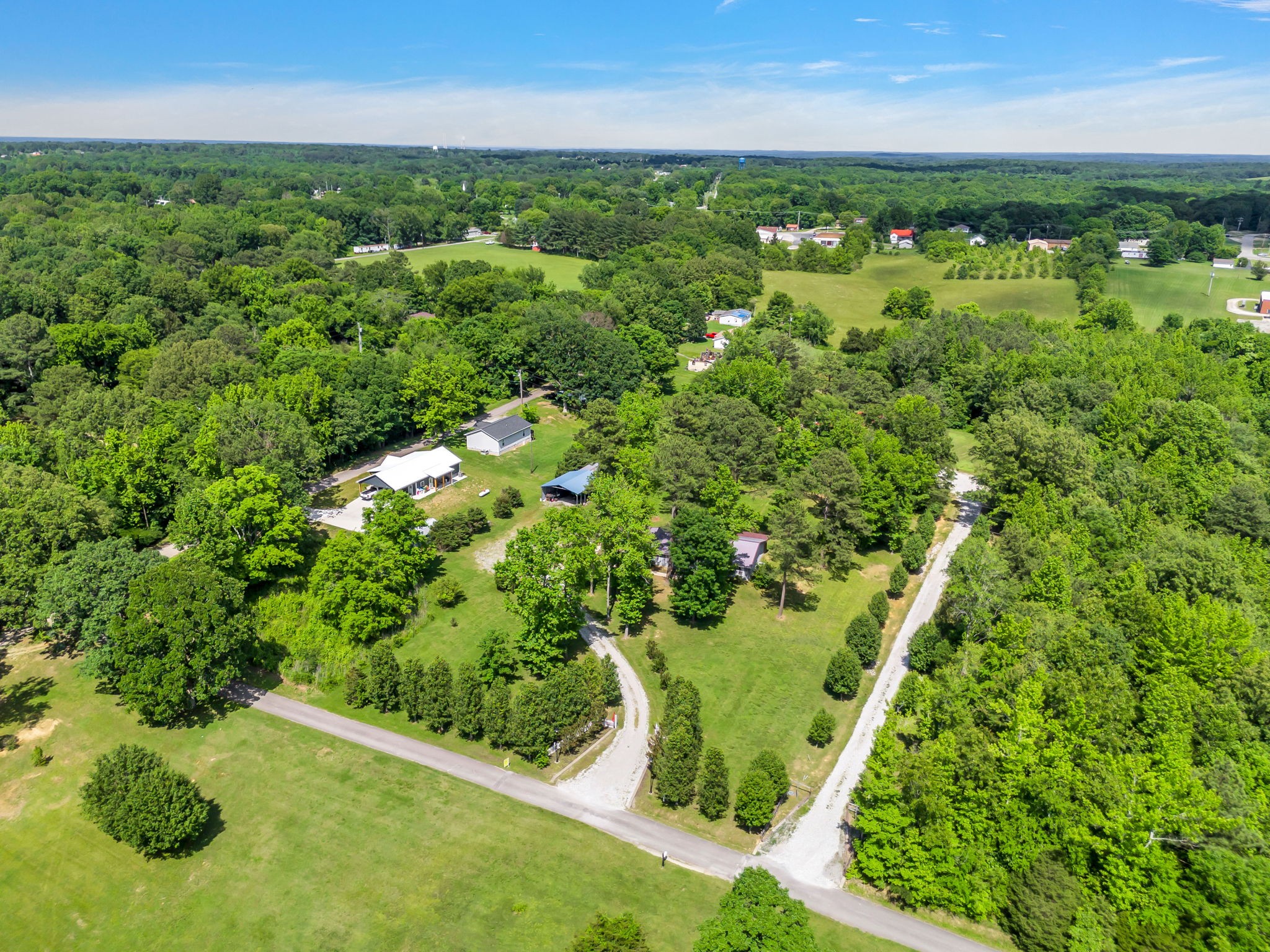 Aerial view of a home and outbuildings amid wooded acreage, open lawns, and gravel drives.