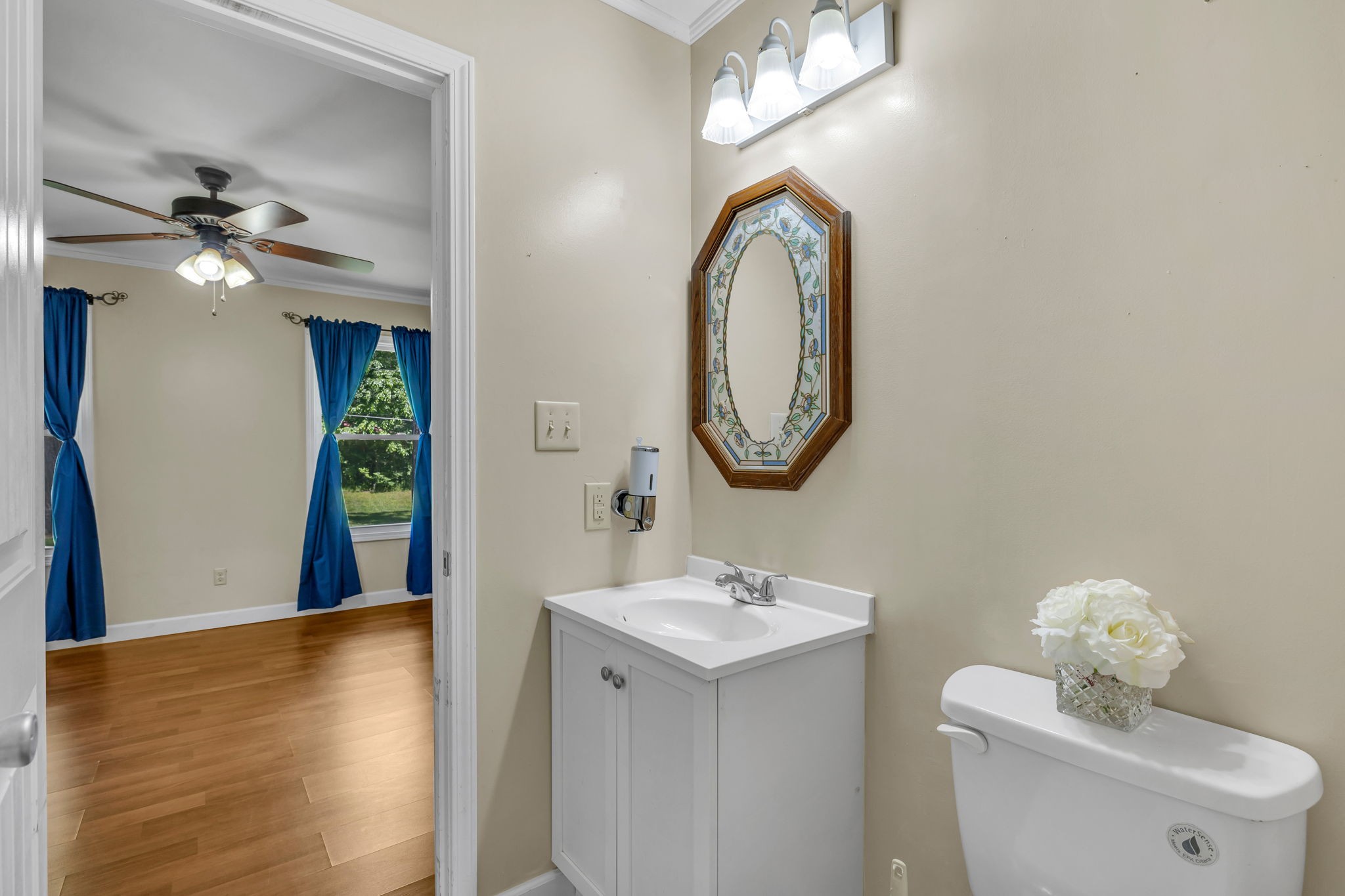 Bathroom with a white vanity, toilet, and octagonal mirror, opening onto a room with blue curtains.