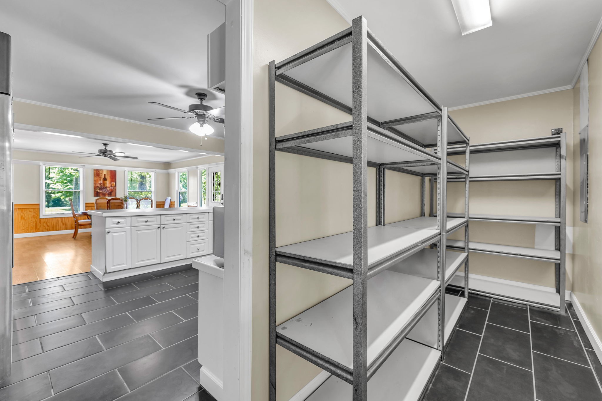 Walk-in pantry with empty metal shelving and dark tile floors beside a bright kitchen.