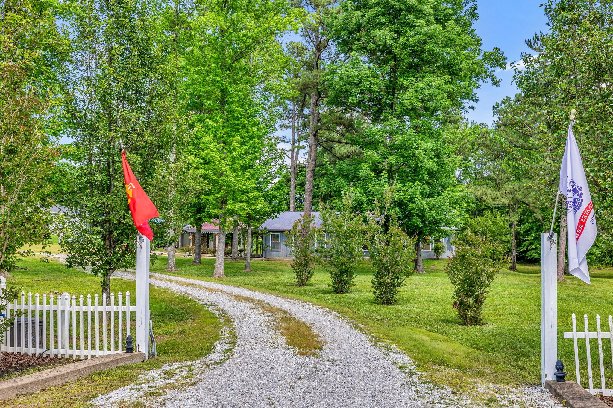 Gray single-story home among mature trees, approached by a curved gravel driveway between white fences.