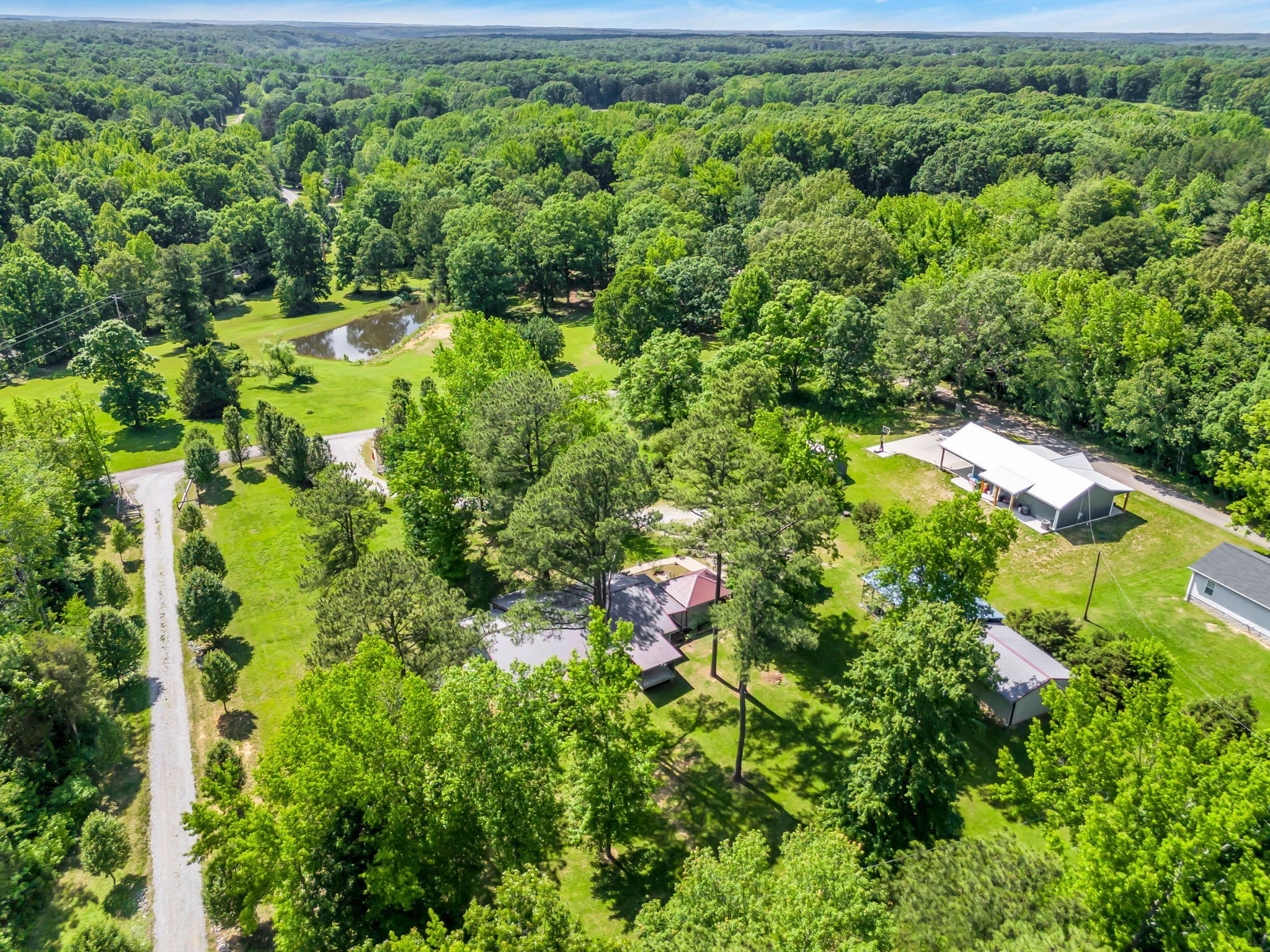 Aerial view of a wooded property with a pond, open lawns, outbuildings, and a gravel driveway.