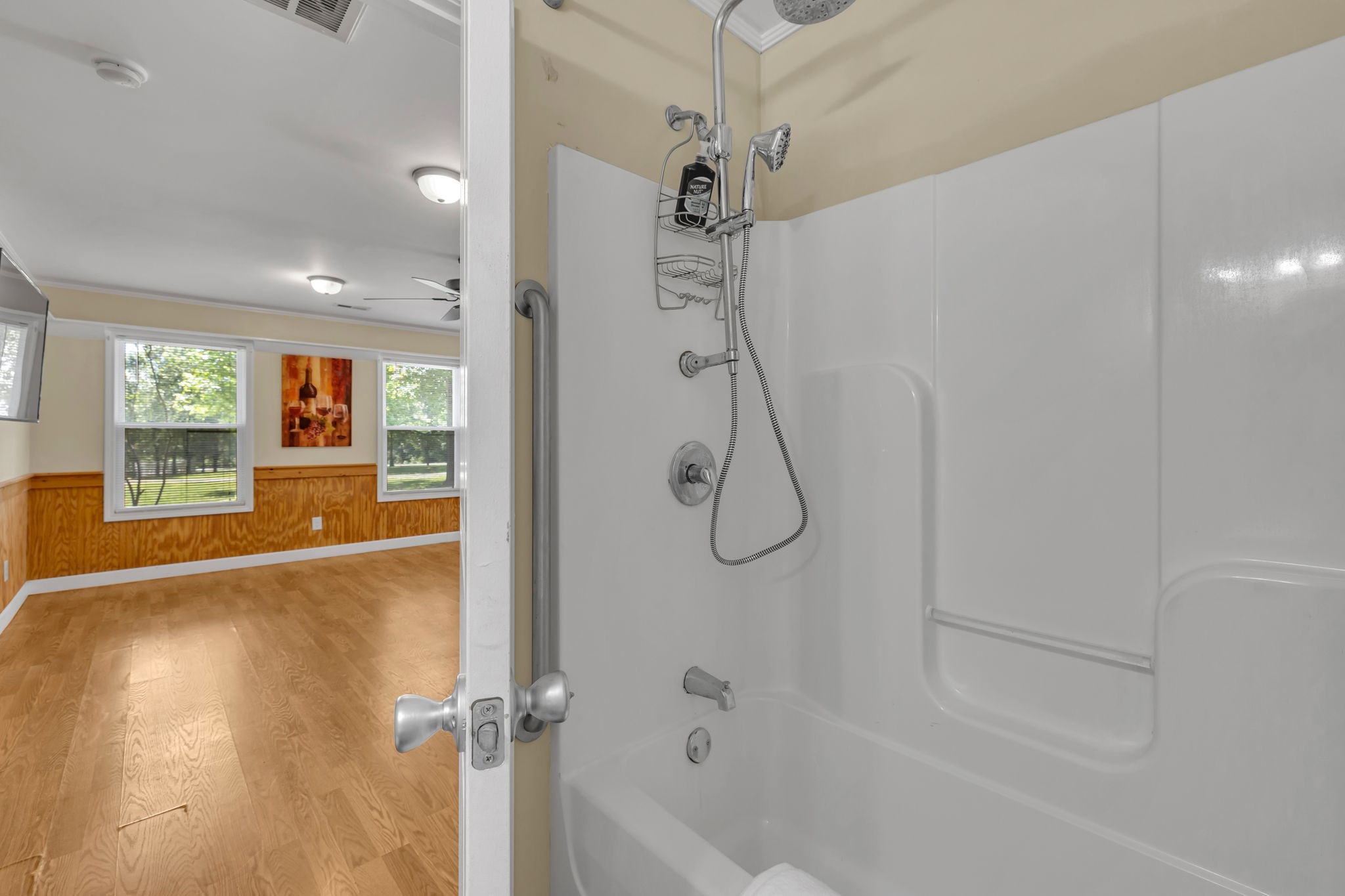 Bathroom with a white tub-shower and handheld showerhead, opening onto a hardwood-floored room.