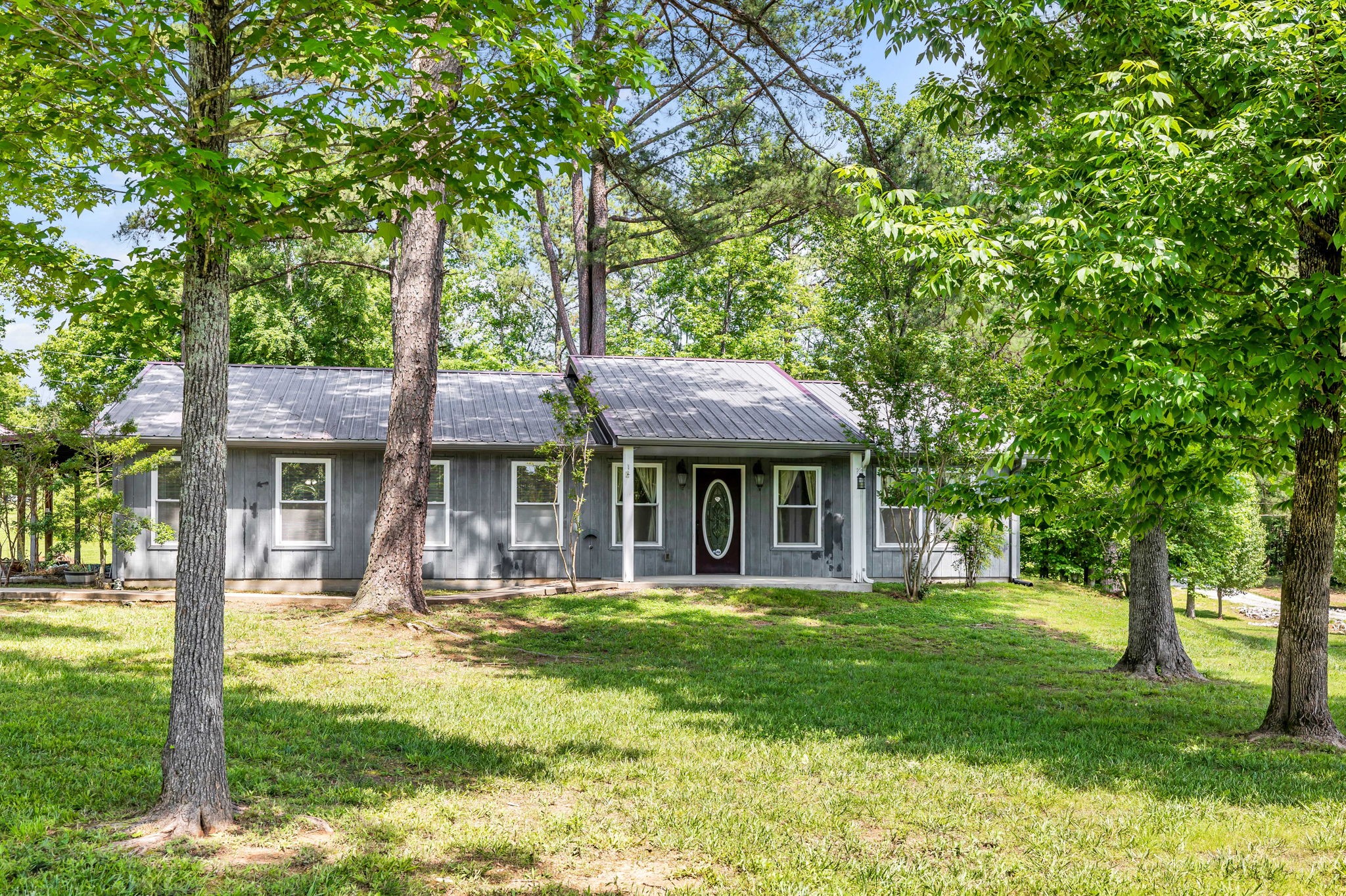 Gray single-story house with a metal roof and oval-glass front door, shaded by mature trees.