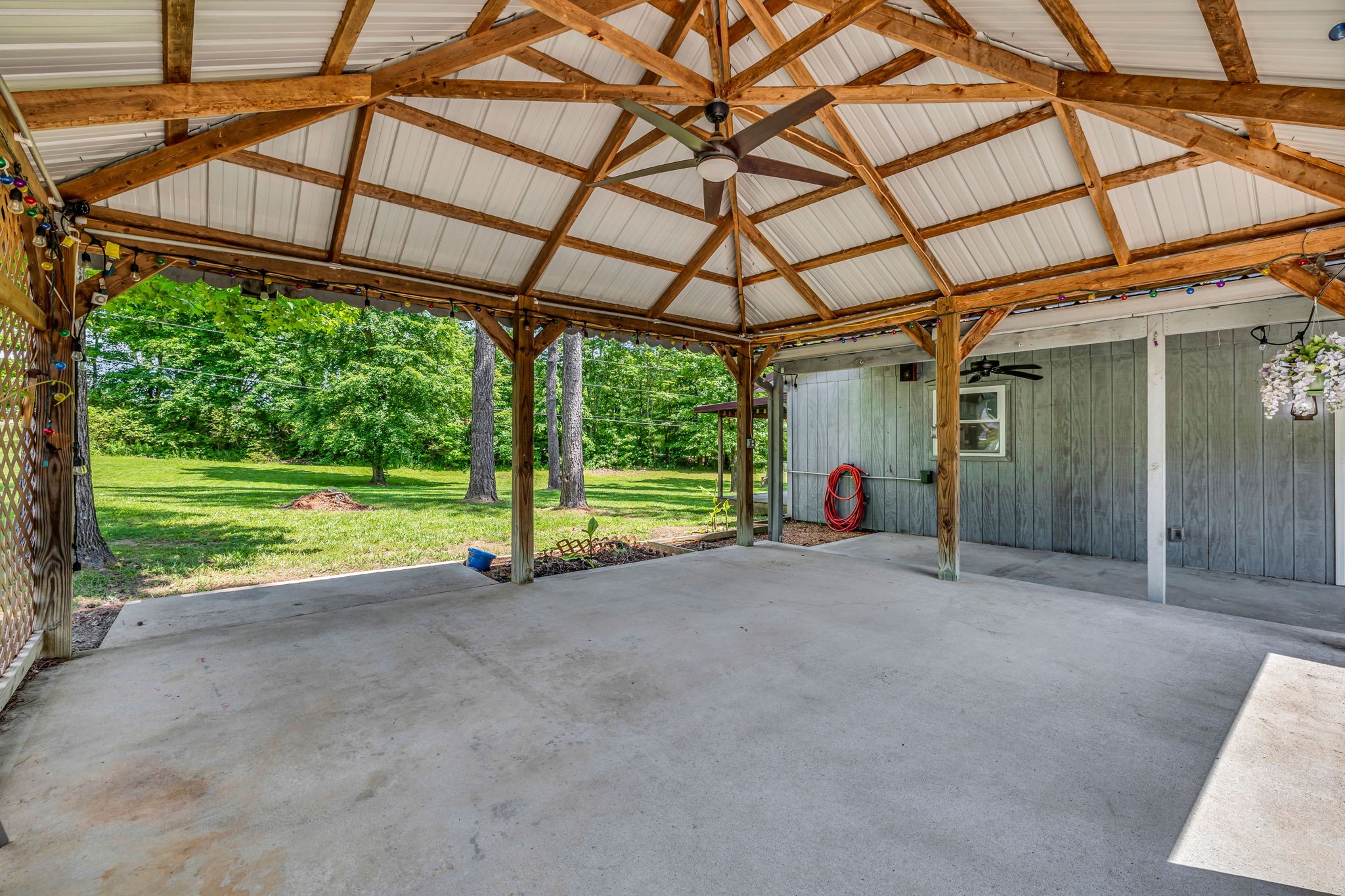 Covered concrete patio with exposed timber trusses, ceiling fan, and a leafy backyard.