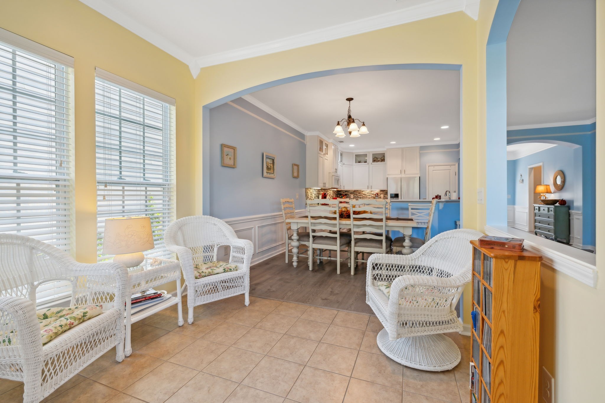 Furnished sitting area with white wicker chairs opening to a dining area and kitchen.