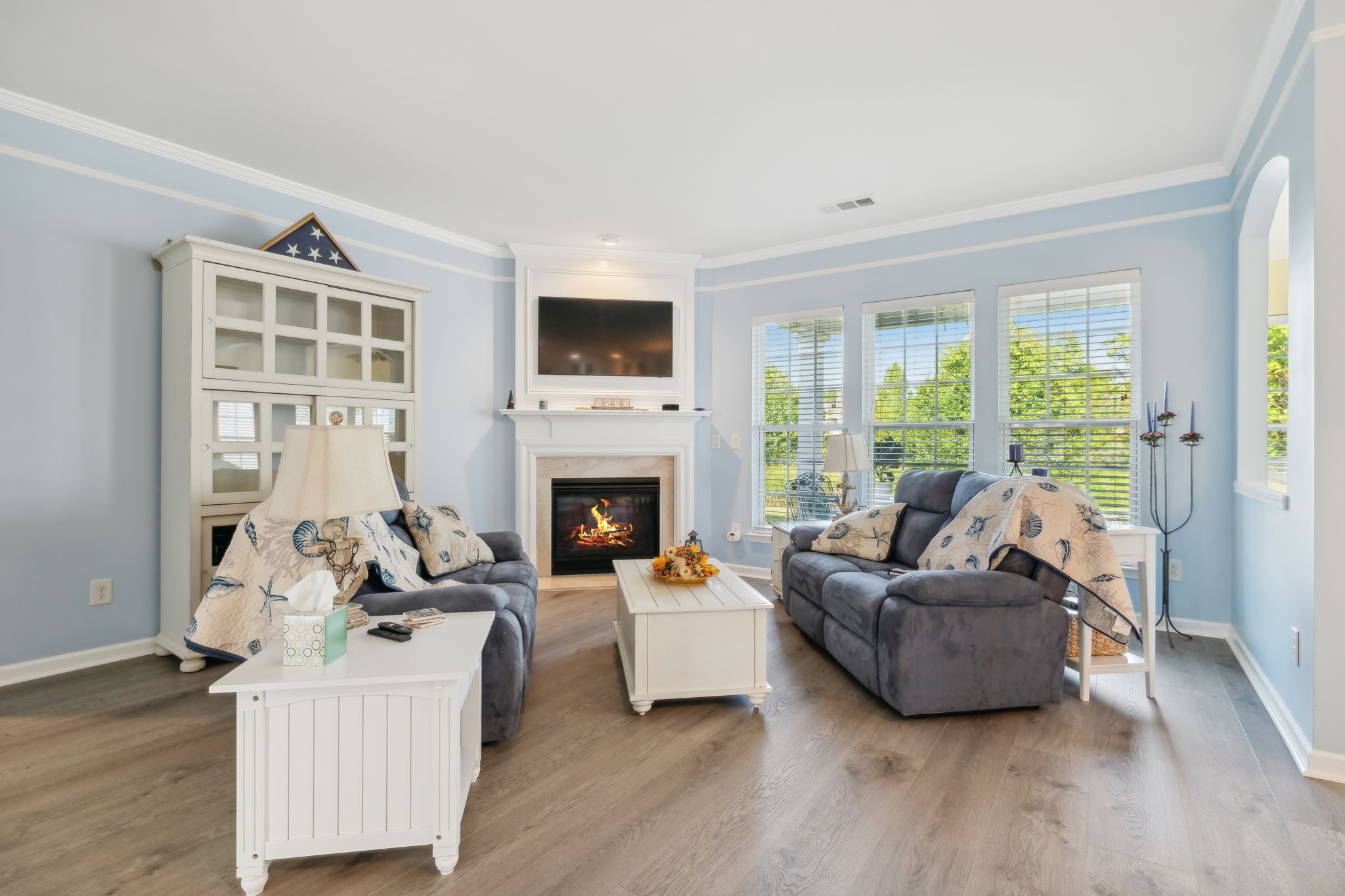 Living room with a lit fireplace, gray sofas, pale blue walls, and large windows overlooking greenery.
