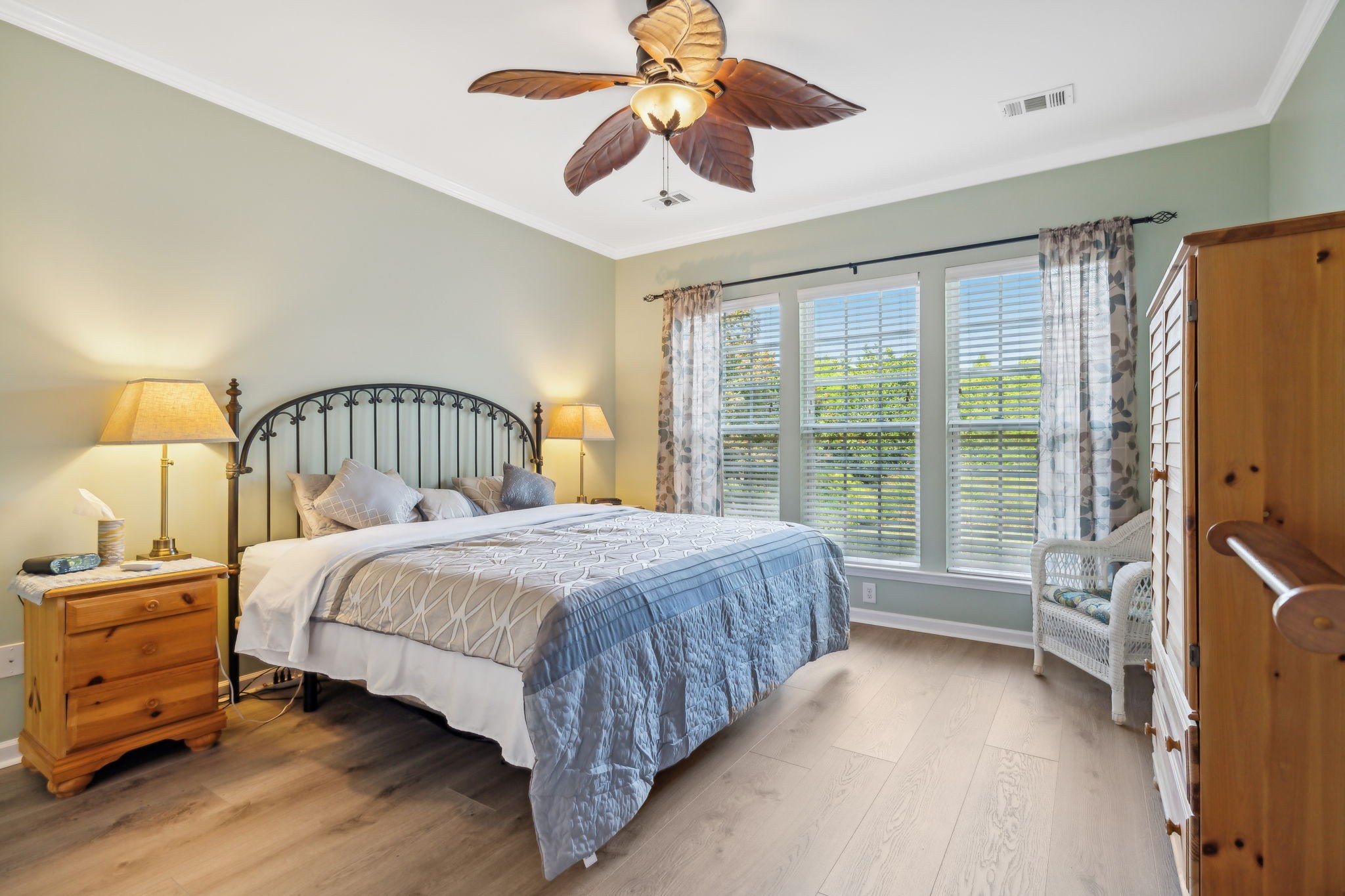 Furnished bedroom with a black metal bed, blue-gray quilt, three curtained windows, and a leaf-shaped ceiling fan.