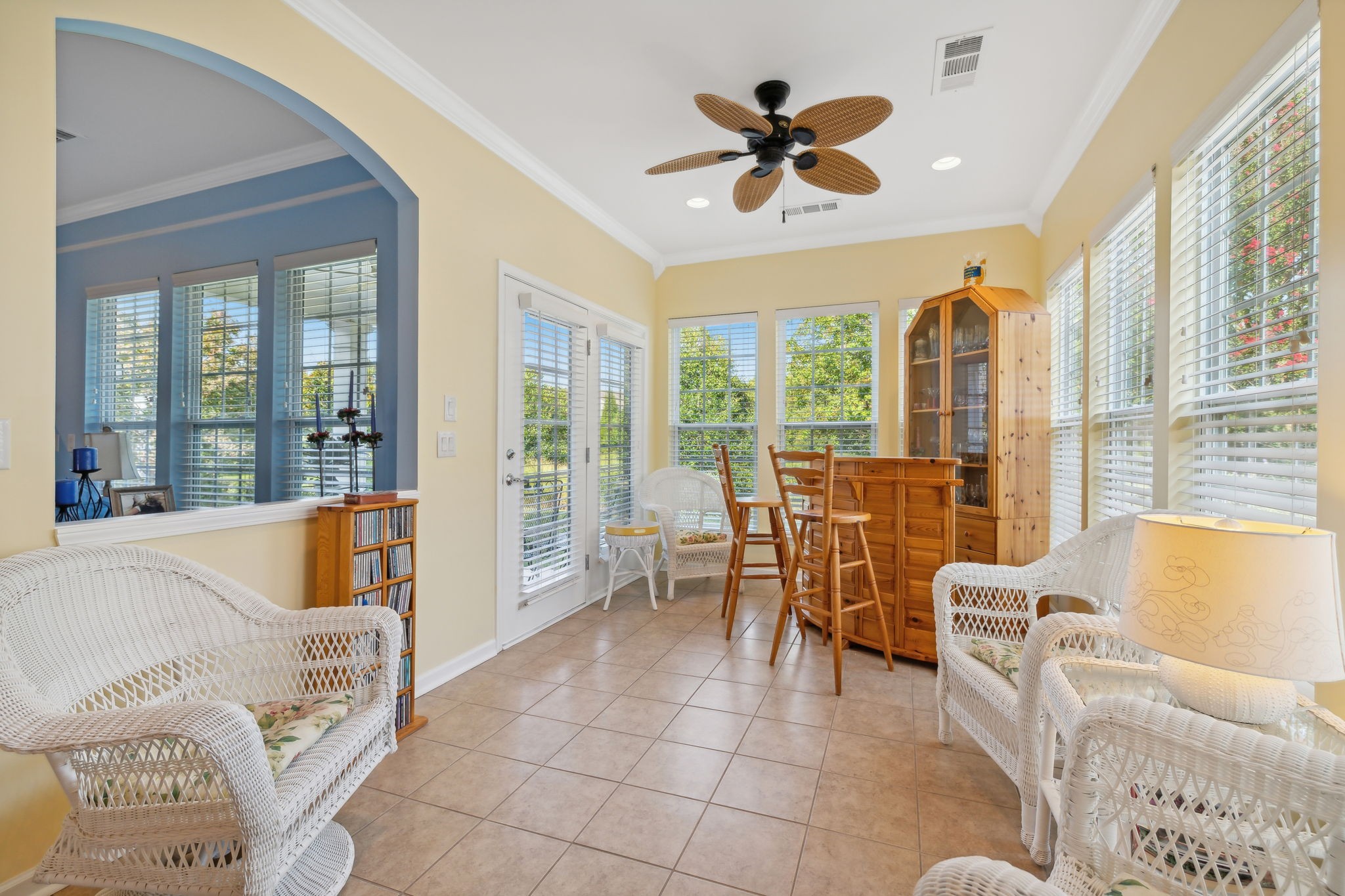 Bright sunroom with wicker seating, tiled floors, and a wooden dining set beside large windows.