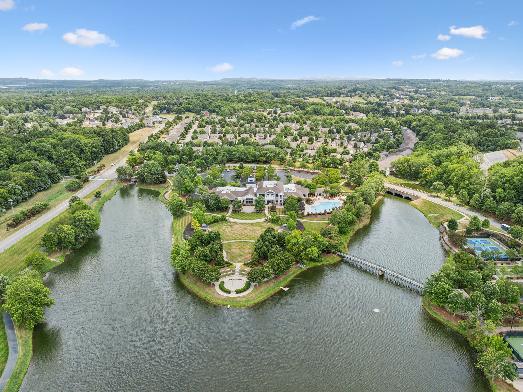 Aerial view of a lakeside clubhouse, pool, walking paths, and tennis courts among homes and trees.