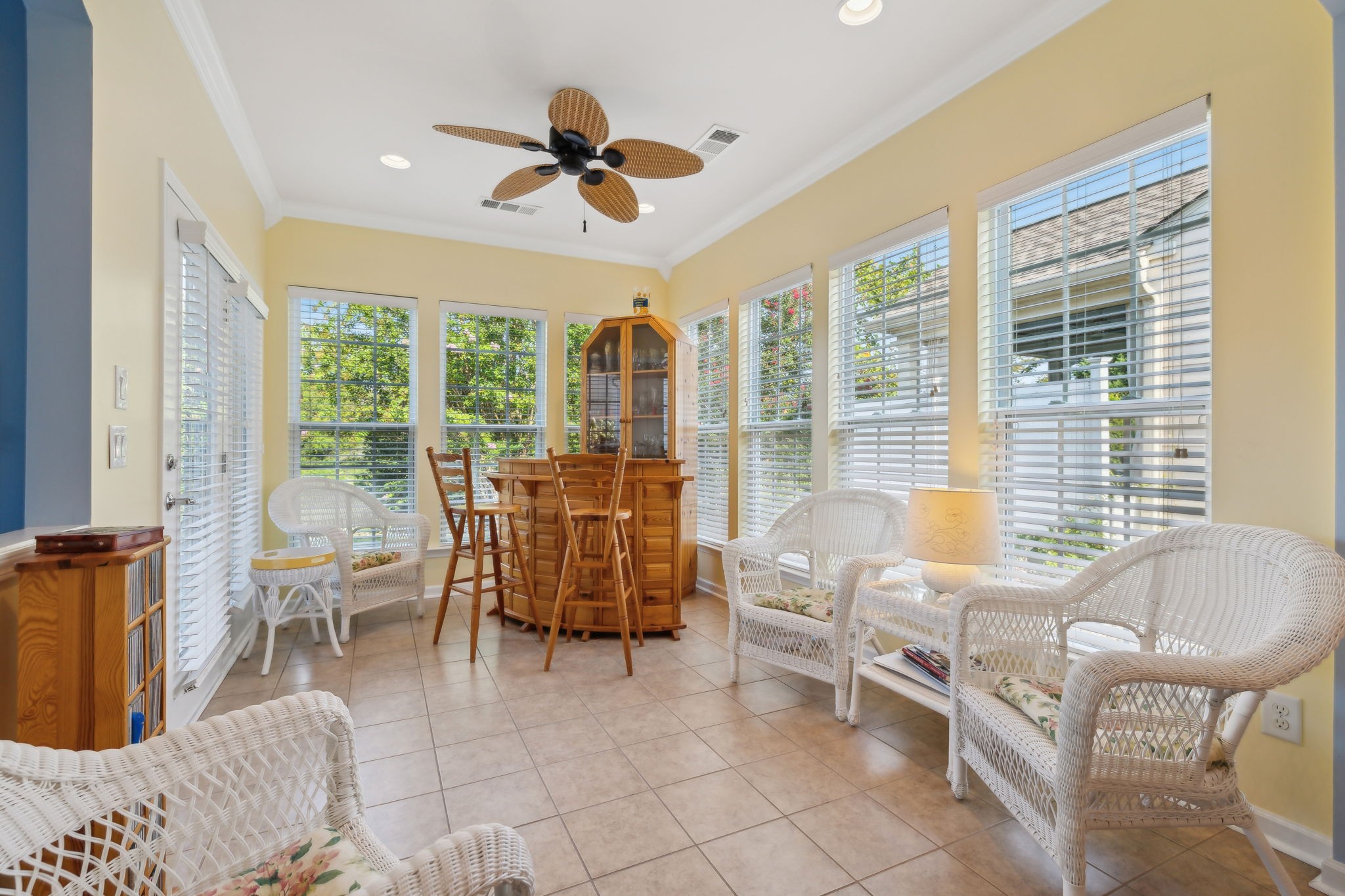 Sunroom with wicker seating, a wooden bar-height table, and large windows overlooking greenery.