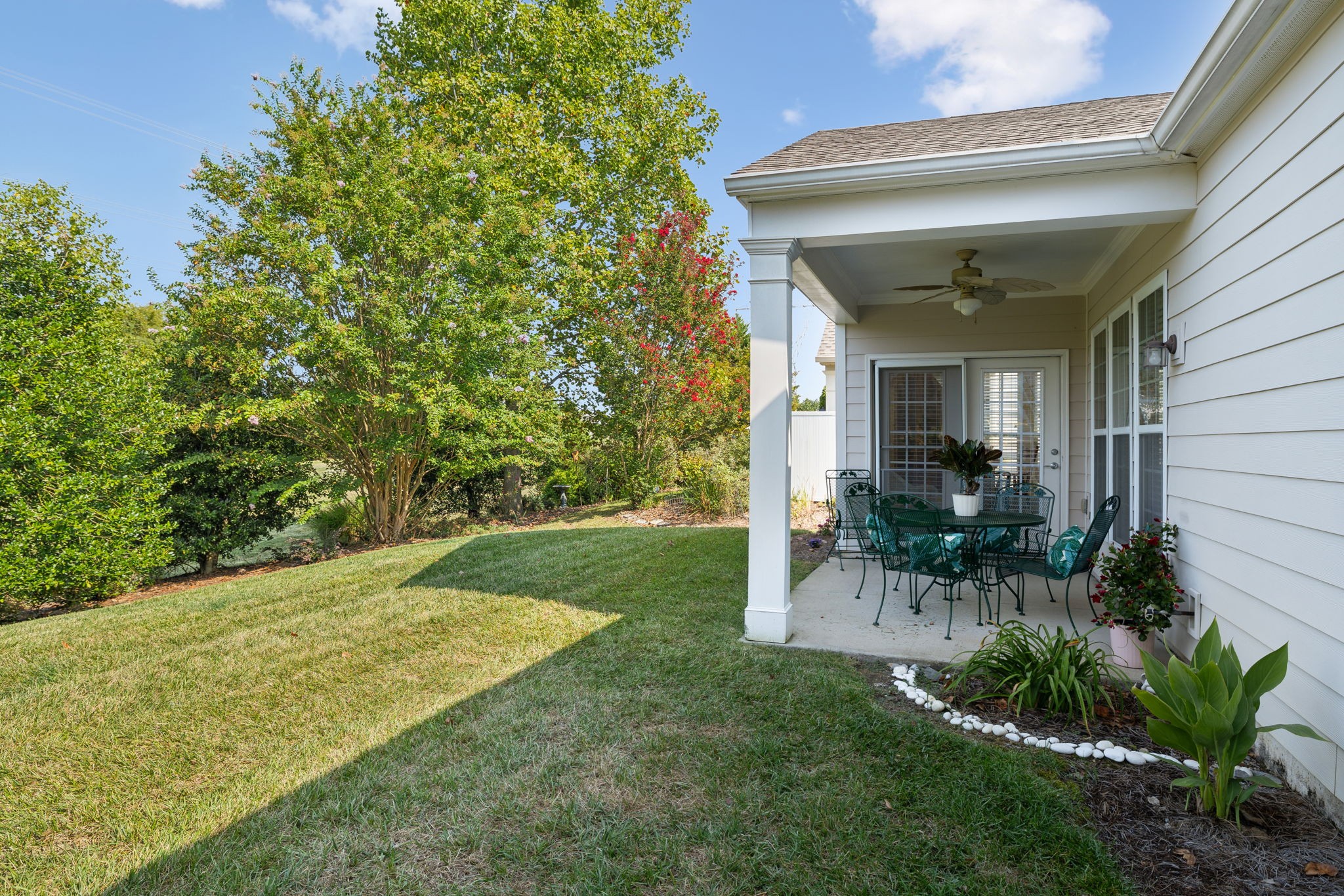 Covered backyard patio with a dining set beside a lawn and mature flowering trees.