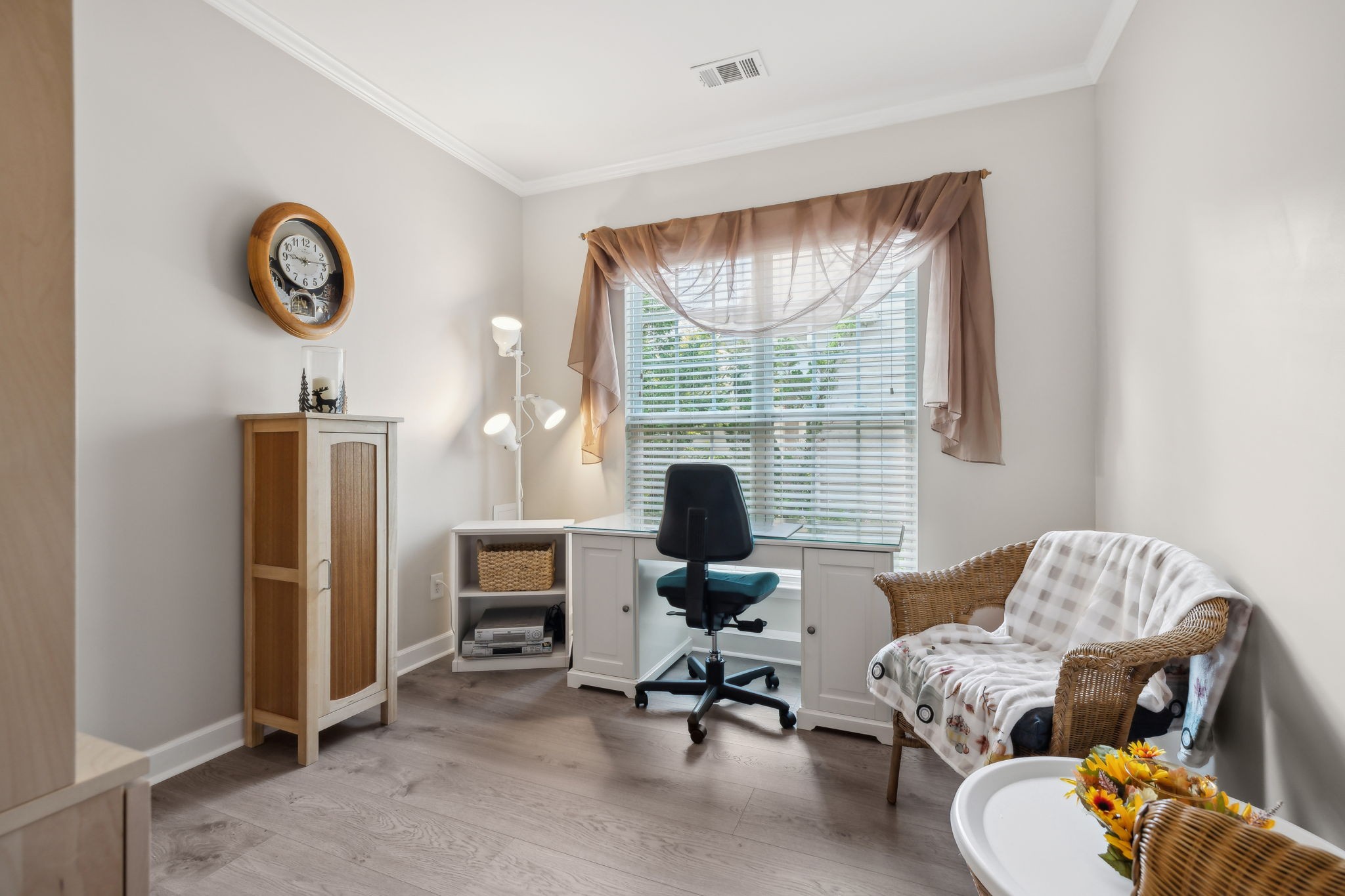 Home office with a white desk beneath a window, black chair, and wicker armchair.