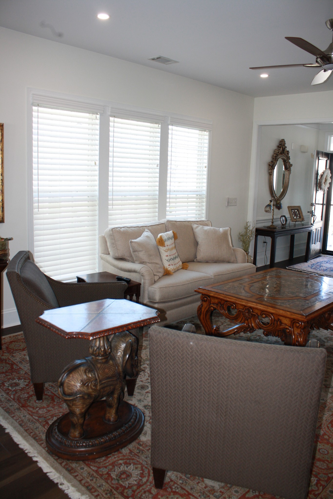Living room with a beige sofa, ornate carved coffee table, and three large windows with blinds.