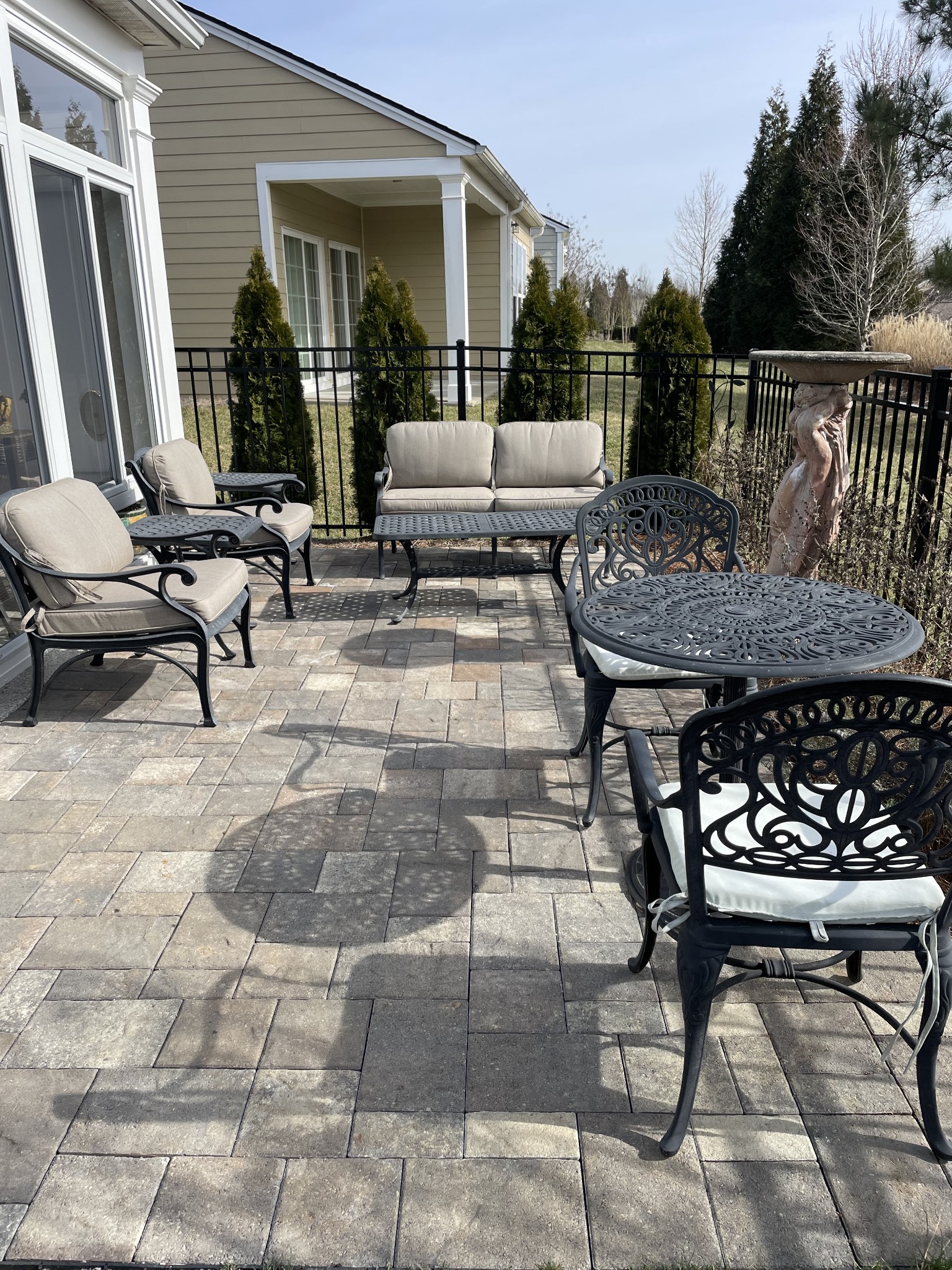 Paved patio with cushioned metal seating, round table, black railing, and birdbath beside a beige home.